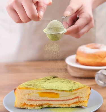 Person dusting matcha powder over a slice of cake with a tea strainer.